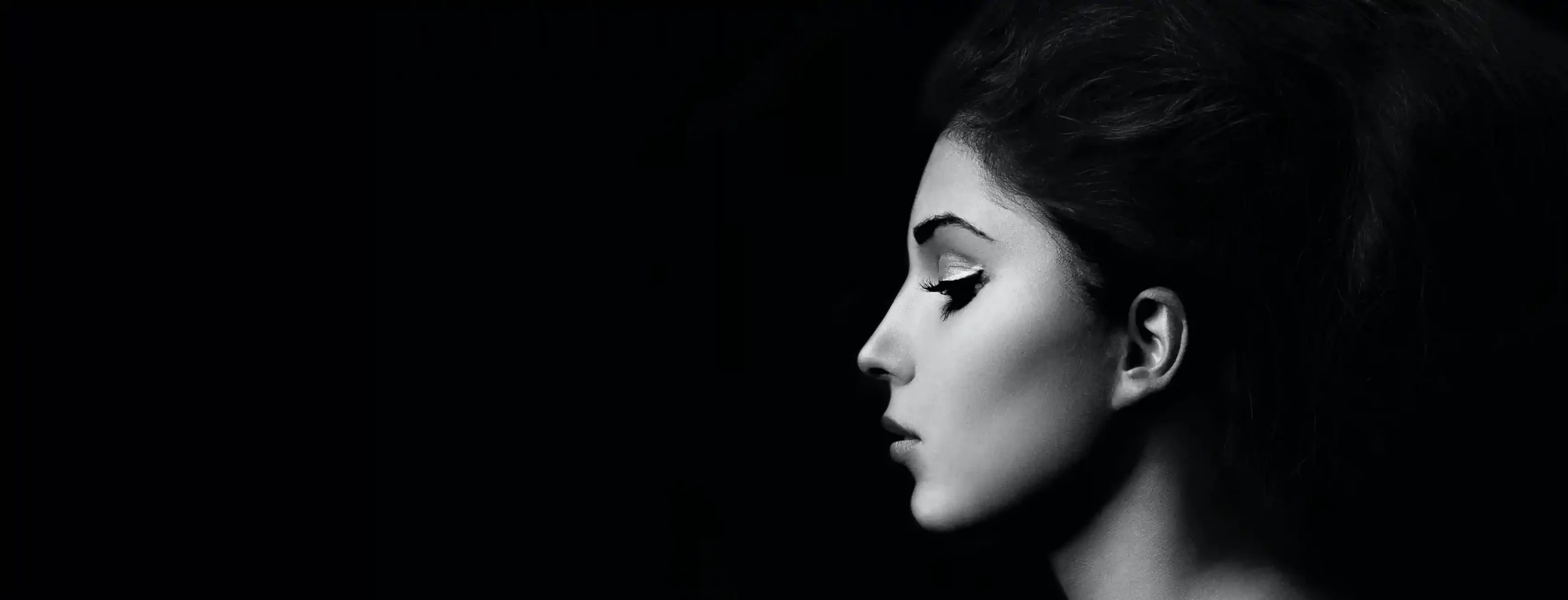 Black and white side profile of a woman with dramatic eye makeup and styled hair against a dark background.