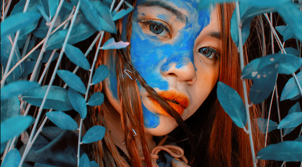 A person with blue face paint, surrounded by green leaves, looks at the camera.