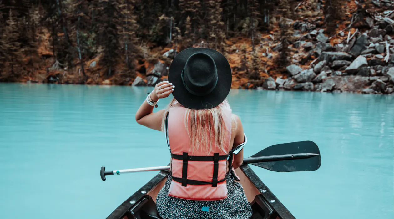 A person wearing a hat and life jacket sits in a canoe holding a paddle, facing turquoise water and a rocky, forested shoreline.