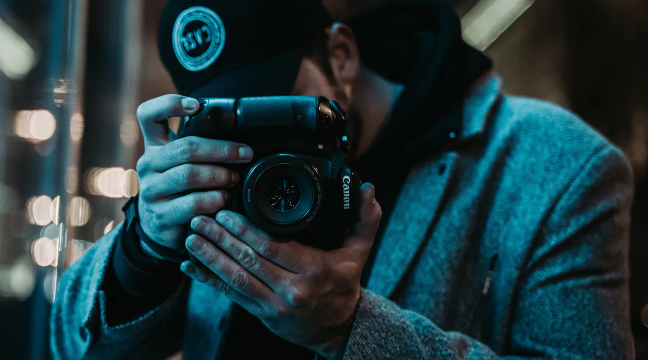 Person wearing a hat and coat holding a camera, taking a photograph in low light.