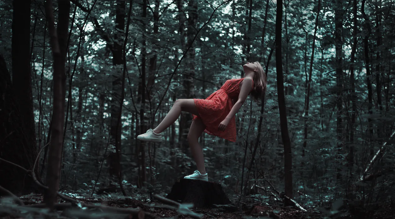 Person in a red dress leans back dramatically on a tree stump in a dark forest.