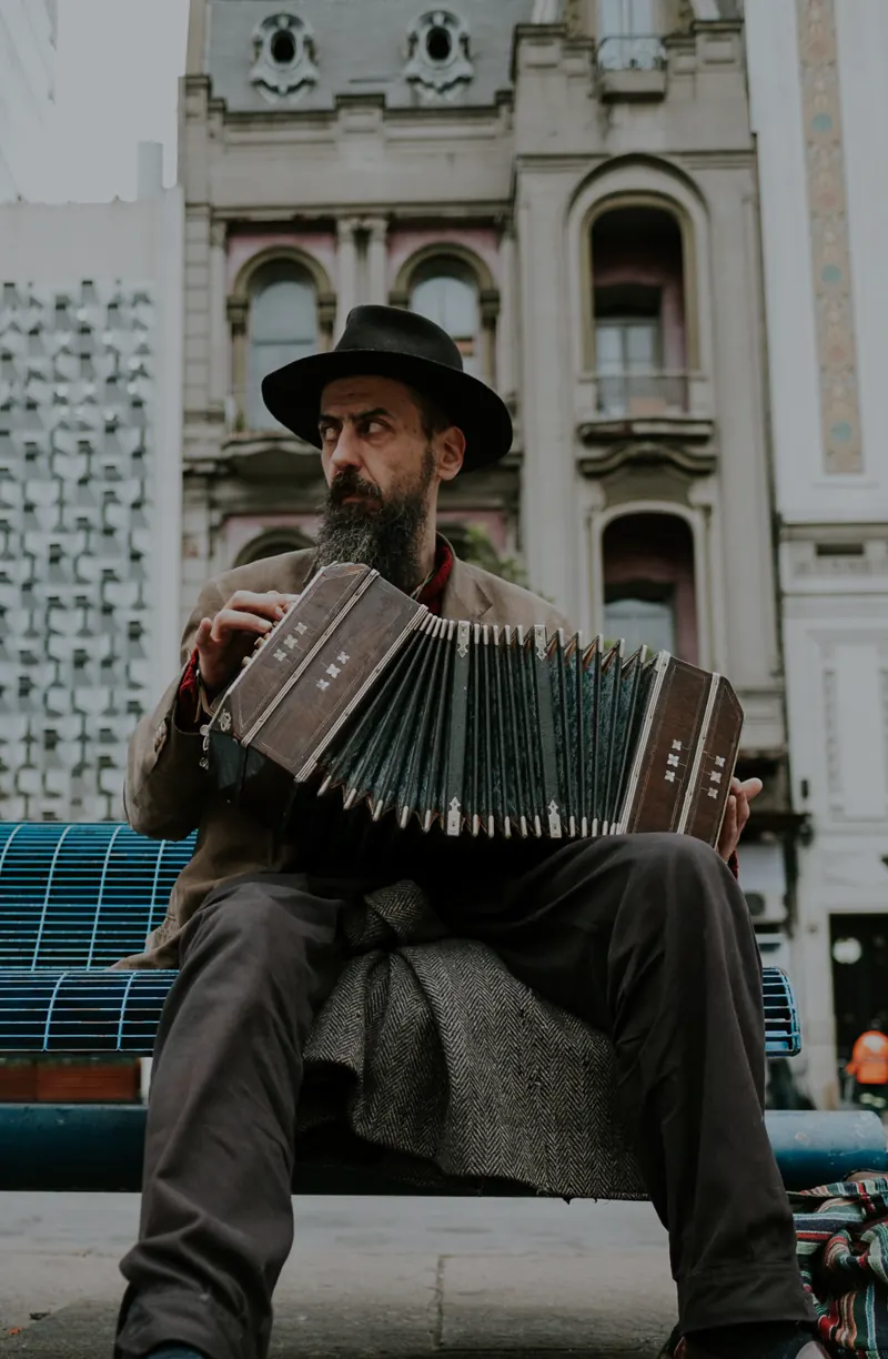 A man with a beard and hat sits on a bench, playing an accordion. Vintage buildings are in the background.