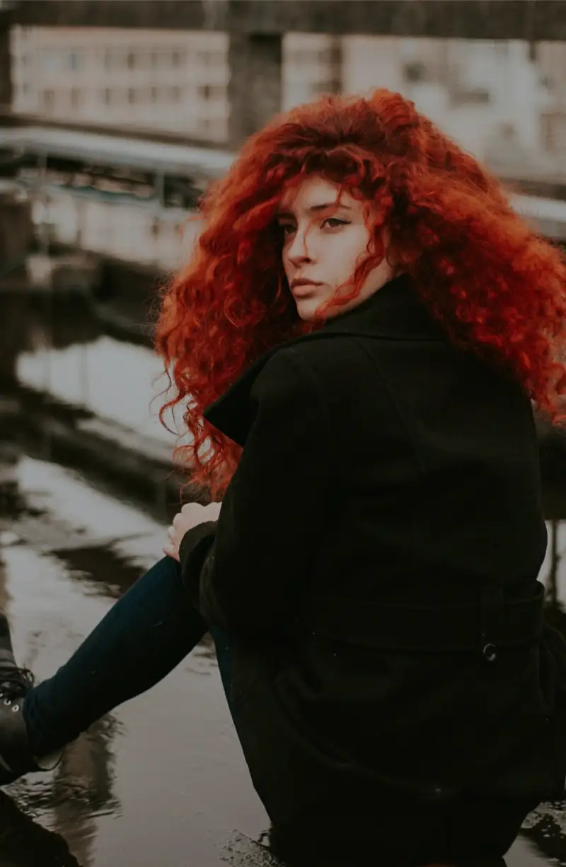 Person with long, curly red hair wearing a black coat sits on a rooftop, looking over their shoulder. Background shows an urban setting with buildings and a gray sky.