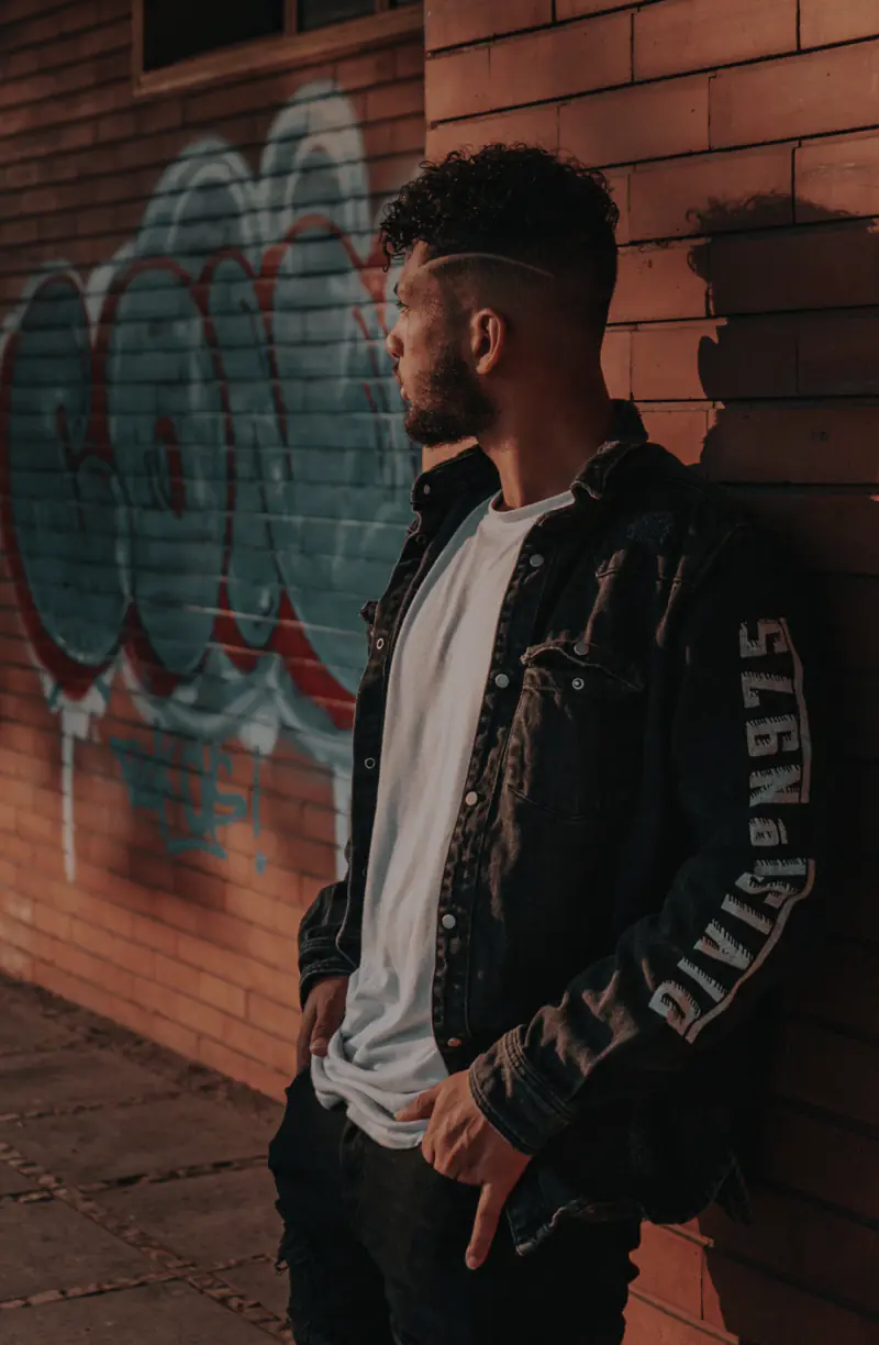 A man with curly hair, wearing a white shirt and dark jacket, leans against a graffiti-covered brick wall, looking to the side.