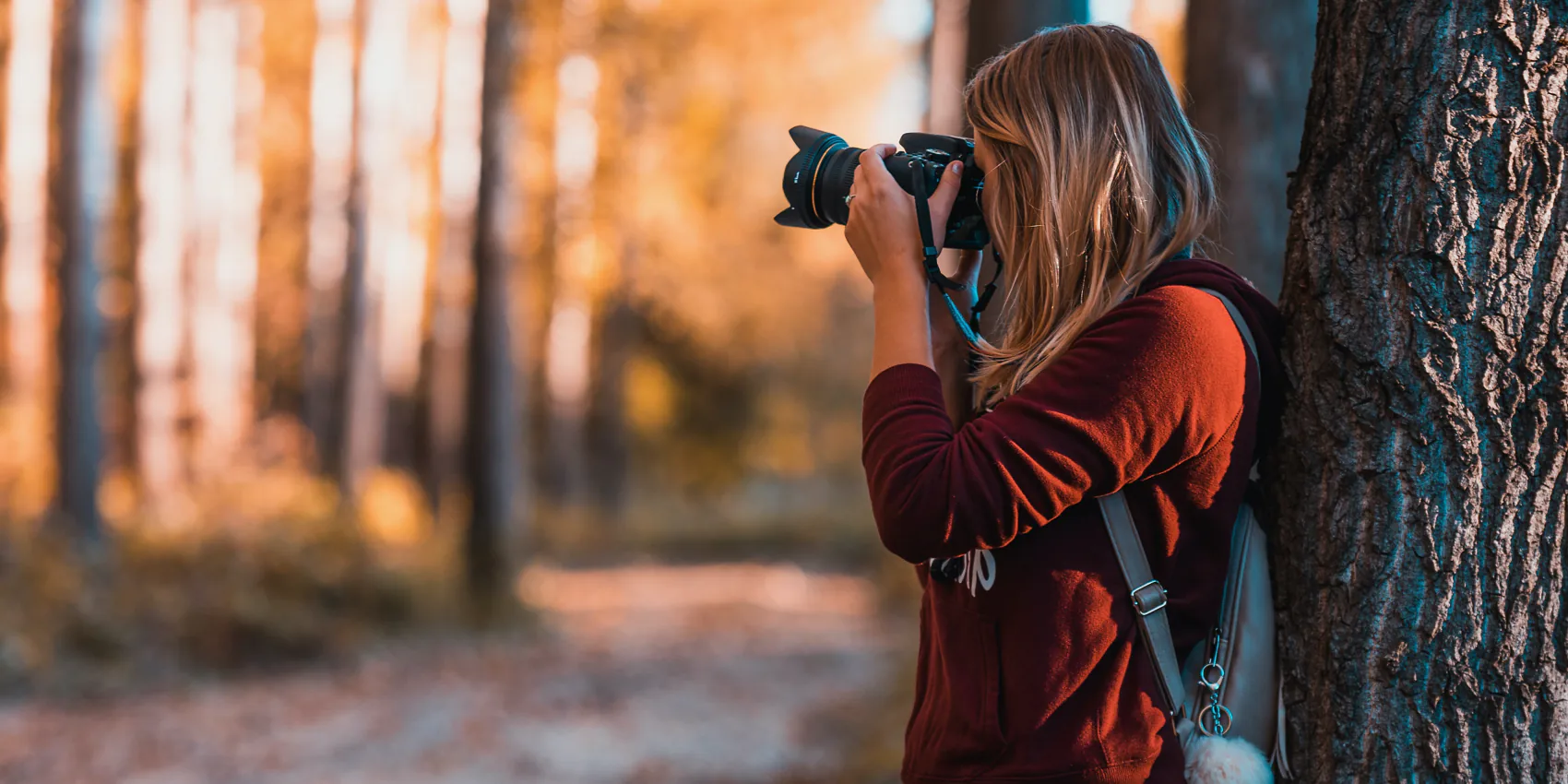 A person with long hair leans against a tree, taking a photo in a forest during autumn.