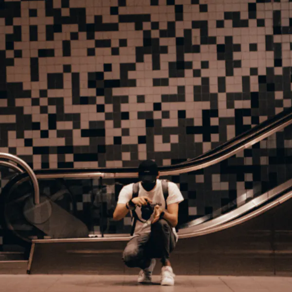 Person kneeling while taking a photo in front of an escalator and a black-and-white checkered wall.