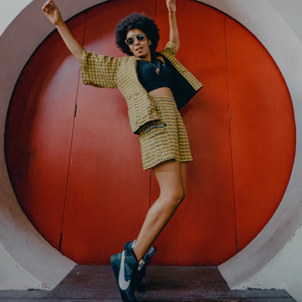 Person in a patterned outfit and sunglasses poses energetically in front of a large red circular door.