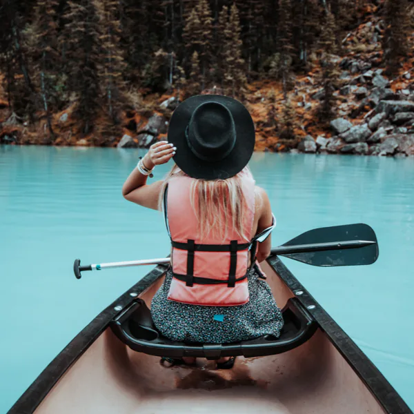 Person in a pink life jacket and black hat paddling a canoe on a turquoise lake, surrounded by trees.