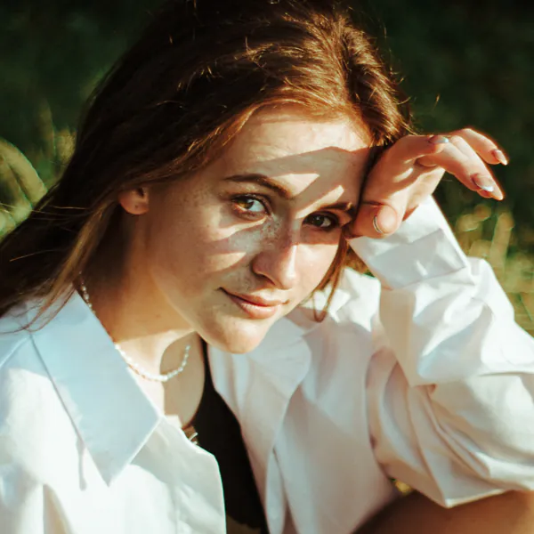 A person with long hair and freckles, wearing a white shirt, sits outdoors with sunlight casting shadows on their face.