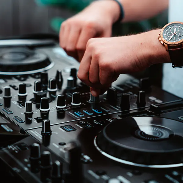 Person adjusting controls on a DJ mixer with both hands, wearing a brown watch on the left wrist.