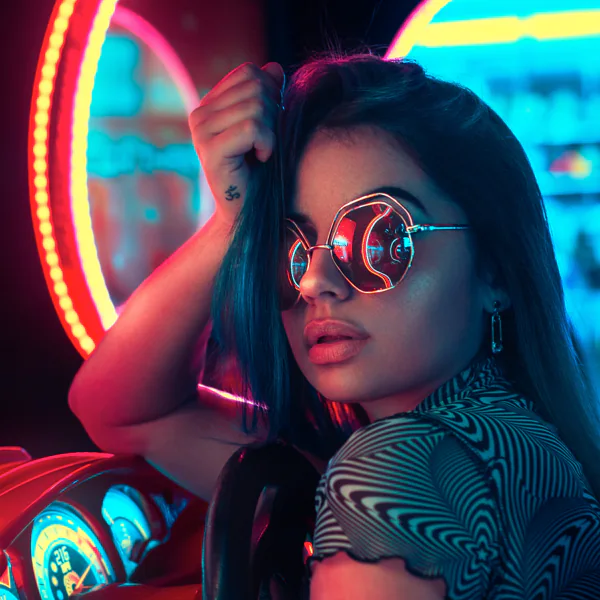 A woman with long hair and round sunglasses poses in a neon-lit arcade, surrounded by colorful lights and gaming machines.