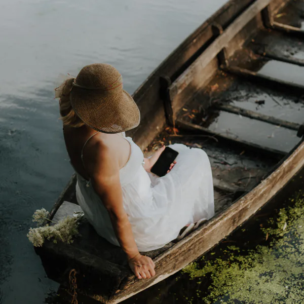 A woman in a white dress and straw hat sits in a wooden boat on a calm lake, holding a smartphone.