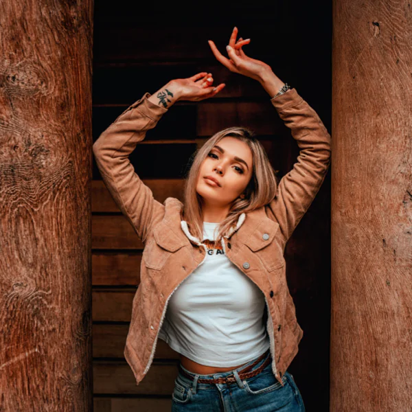 Person in a tan jacket and jeans poses with arms raised between two wooden posts.