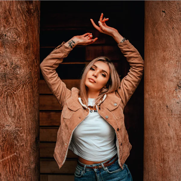 A person in a tan jacket and jeans poses with arms raised between two wooden pillars.