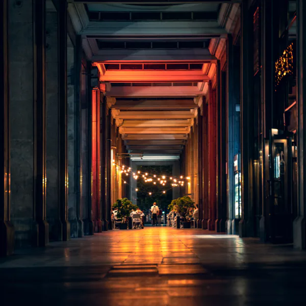 A corridor with illuminated string lights and columns, leading to a well-lit area with tables and people at the end.