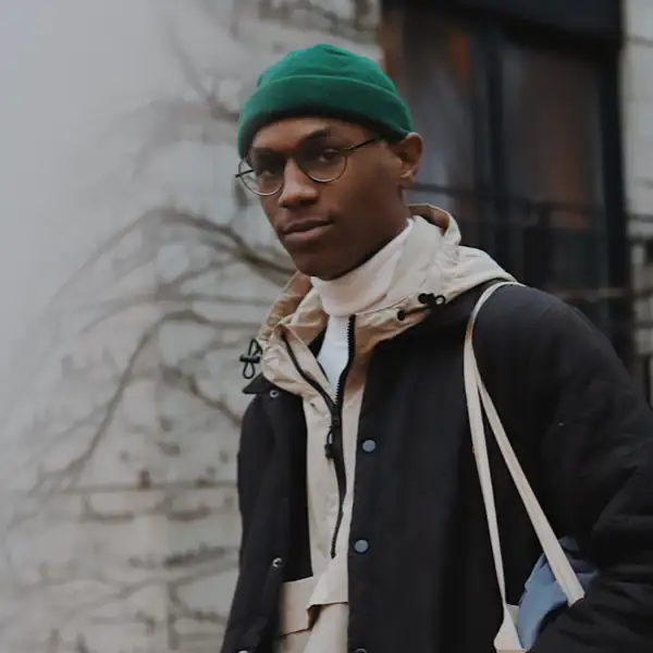 A person wearing glasses, a green beanie, and layered jackets stands outside near a building.