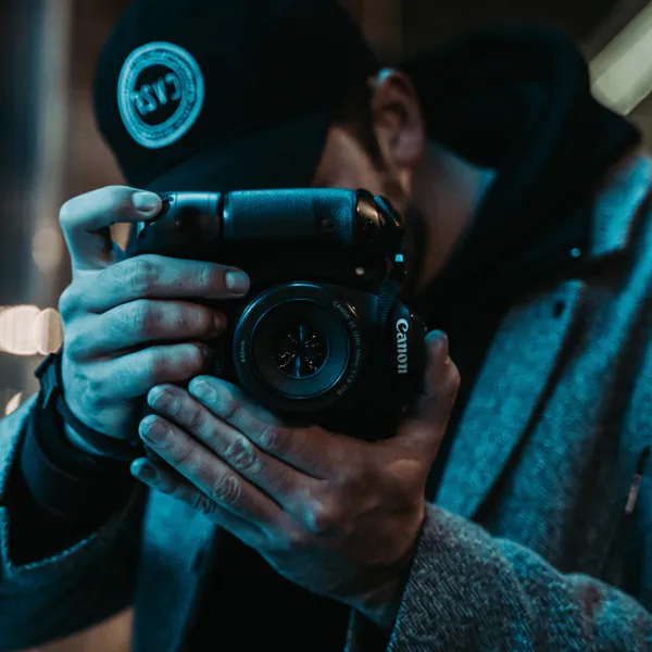 A person wearing a cap and coat holds a Canon camera, focusing on taking a photo in a dimly lit environment.