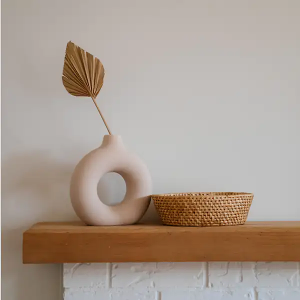 A round vase with a dried leaf and a woven basket sit on a wooden mantel against a beige wall.