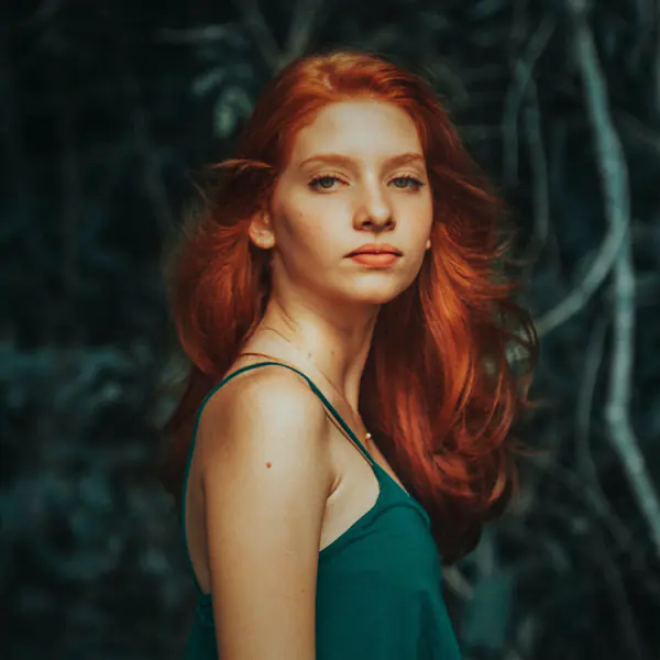 A person with long red hair stands wearing a teal top, with a background of dark greenery.