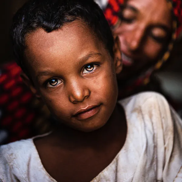 Child with serious expression in foreground, wearing a white shirt. Smiling person in colorful headscarf in soft-focus background.