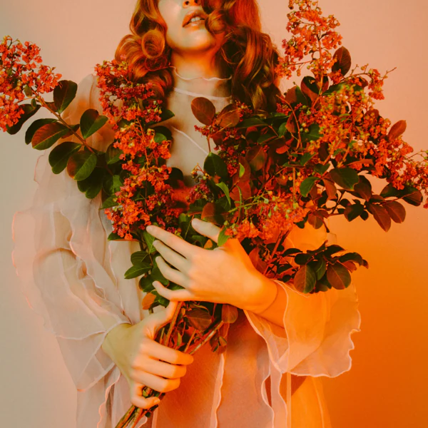 A person with wavy hair, wearing a sheer outfit, is holding a large bouquet of red flowers against a warm, amber-toned background.