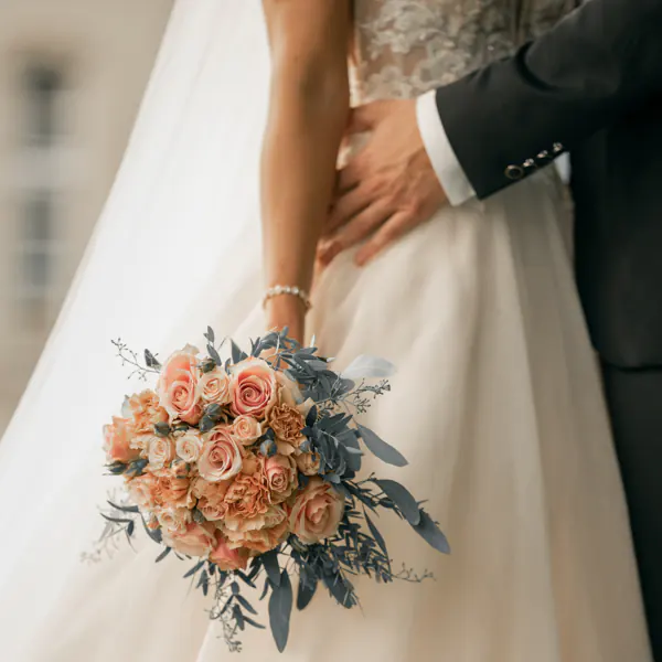 Close-up of a bride holding a bouquet of pink roses and greenery, standing with a groom in a dark suit, with his arm around her waist.