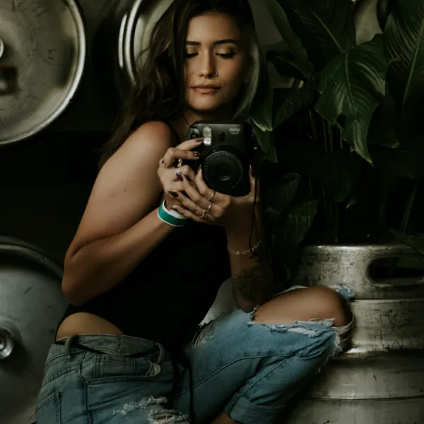 A woman in a black top and ripped jeans is holding a camera, surrounded by metal barrels and green plants.