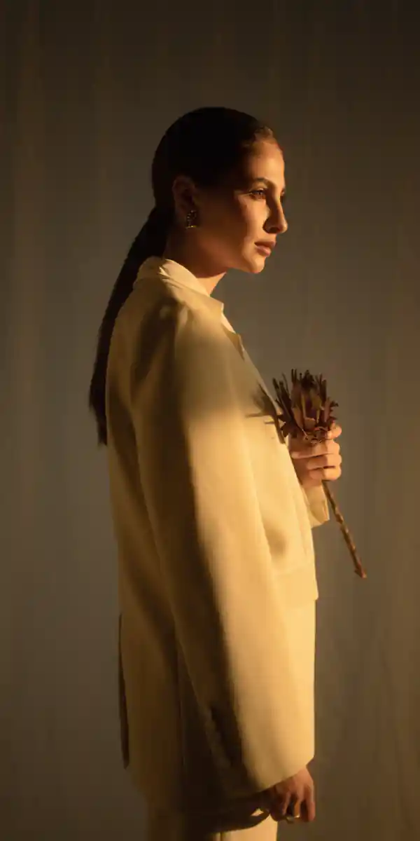 A woman in a beige suit stands holding dried flowers, with a side profile view, illuminated by warm light against a neutral background.