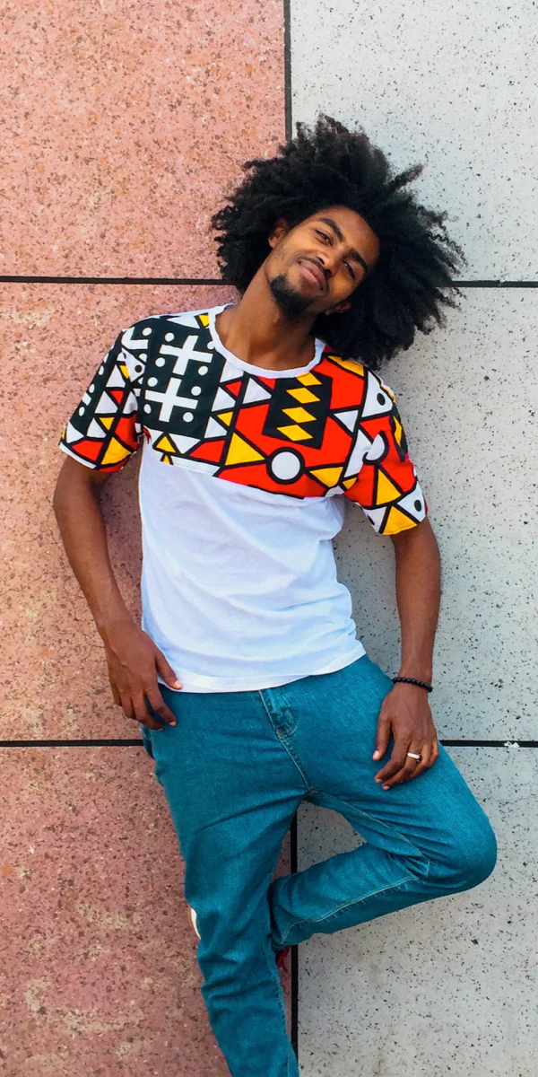 Man with curly hair leans against a tiled wall, wearing a colorful geometric patterned shirt and jeans.