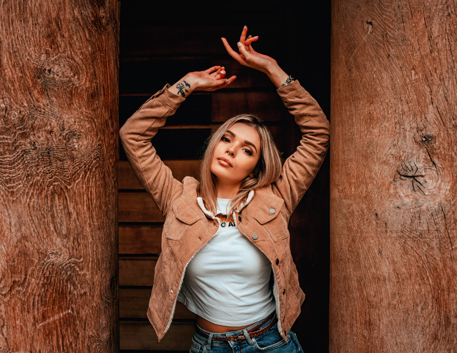 Woman in a tan jacket and white shirt poses with arms raised against a wooden background.