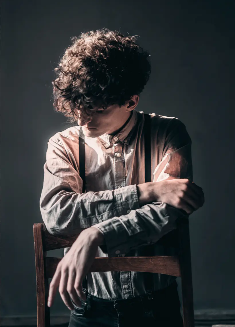 Person with curly hair sits on a wooden chair, looking down, with arms crossed over the chair's backrest. Dramatic lighting creates shadows on a dark background.