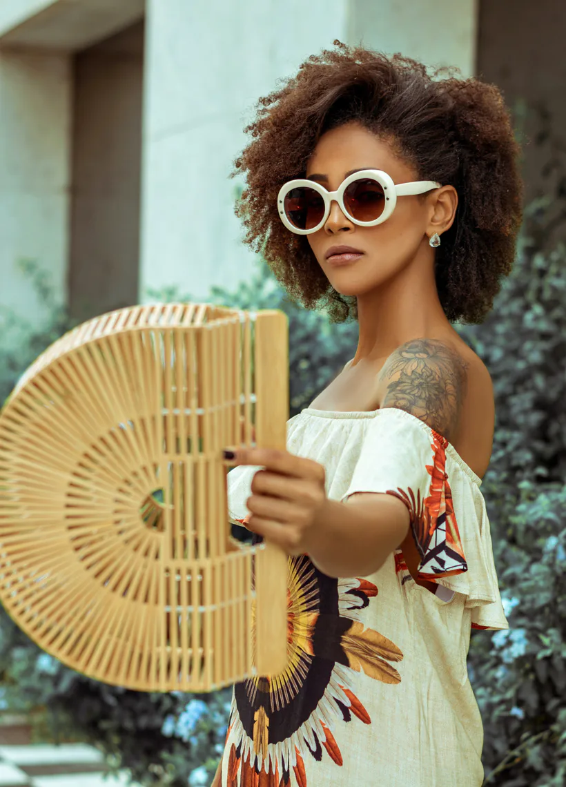 Woman with curly hair and sunglasses holds a wooden fan. She wears a patterned dress and stands outside with greenery in the background.