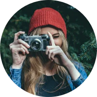 Person wearing a red beanie and blue jacket holding a camera up to their face, standing outdoors with greenery in the background.