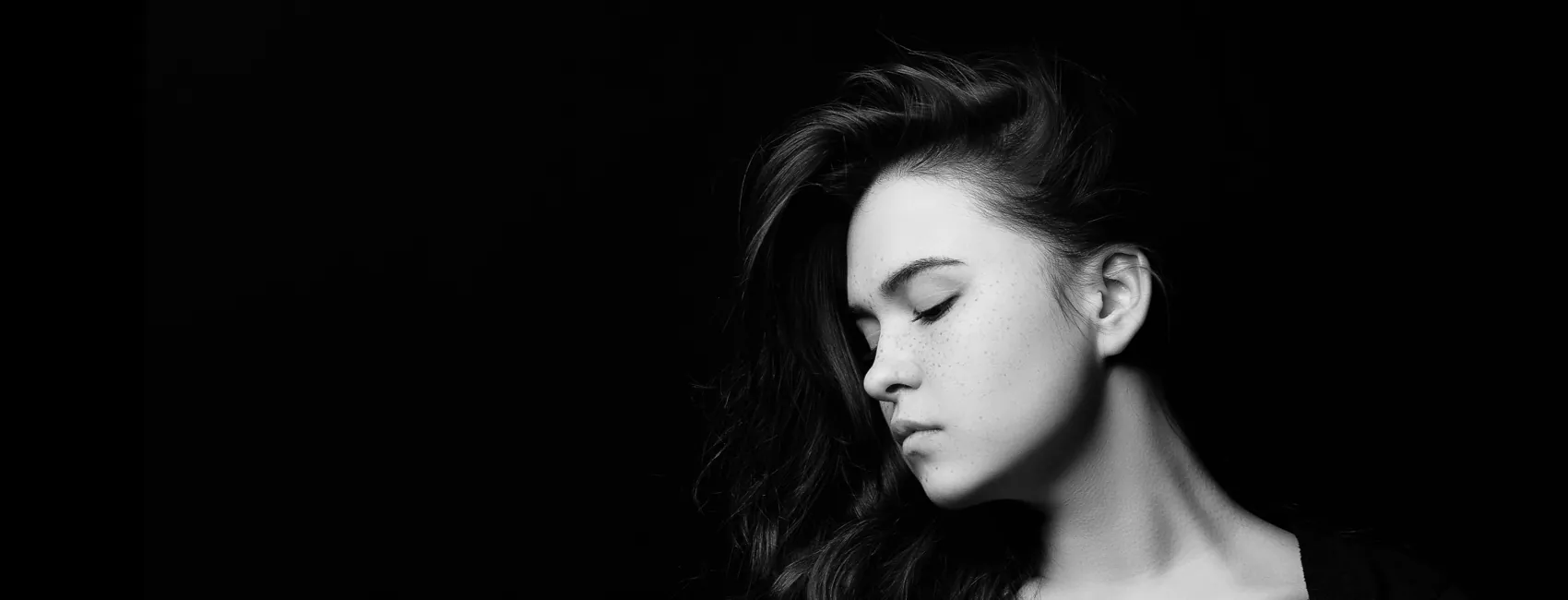 A woman with long dark hair and closed eyes is posed against a black background in a side profile. The image is in black and white.