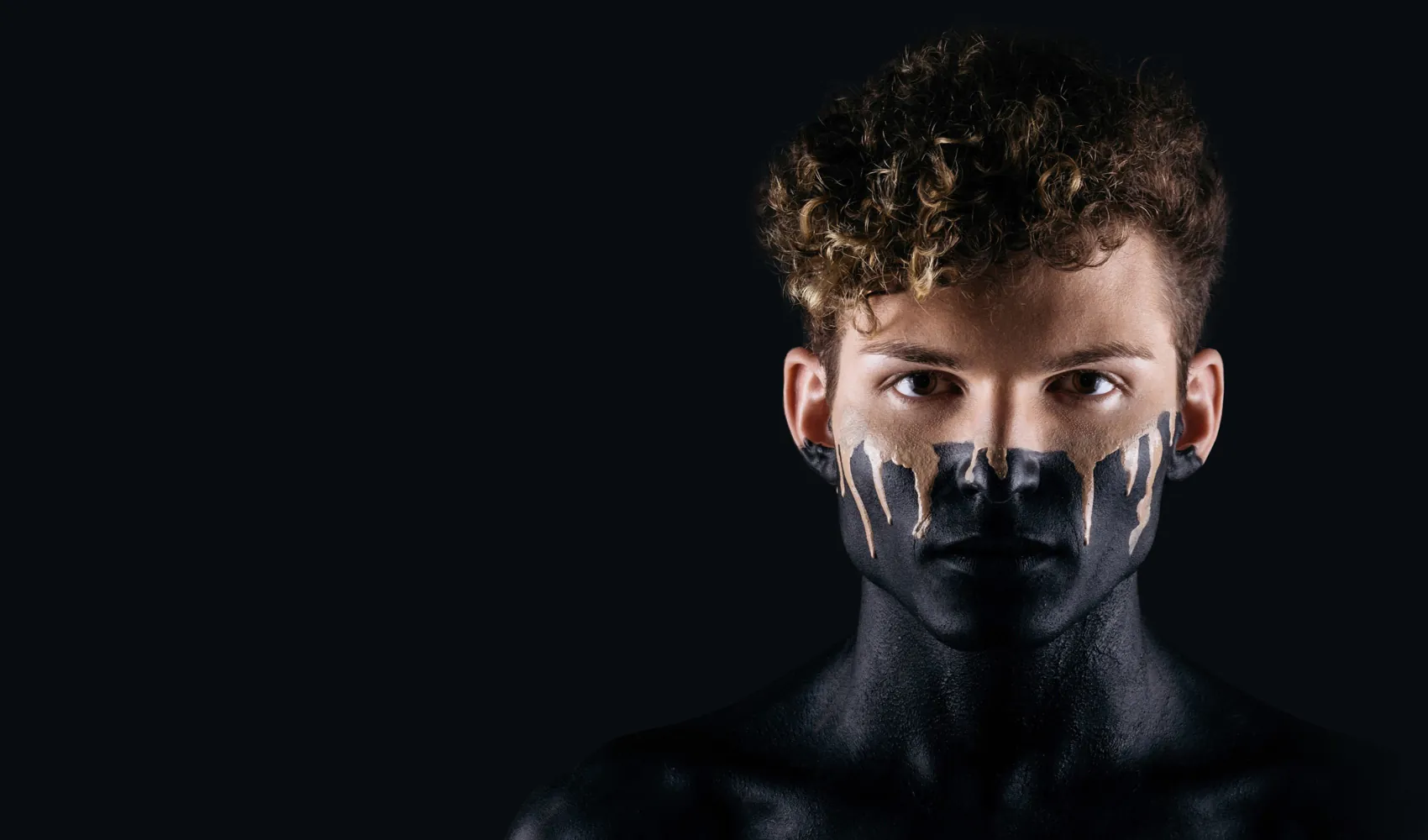 Person with curly hair, face, and upper body painted black, with white paint drips on the face, against a dark background.