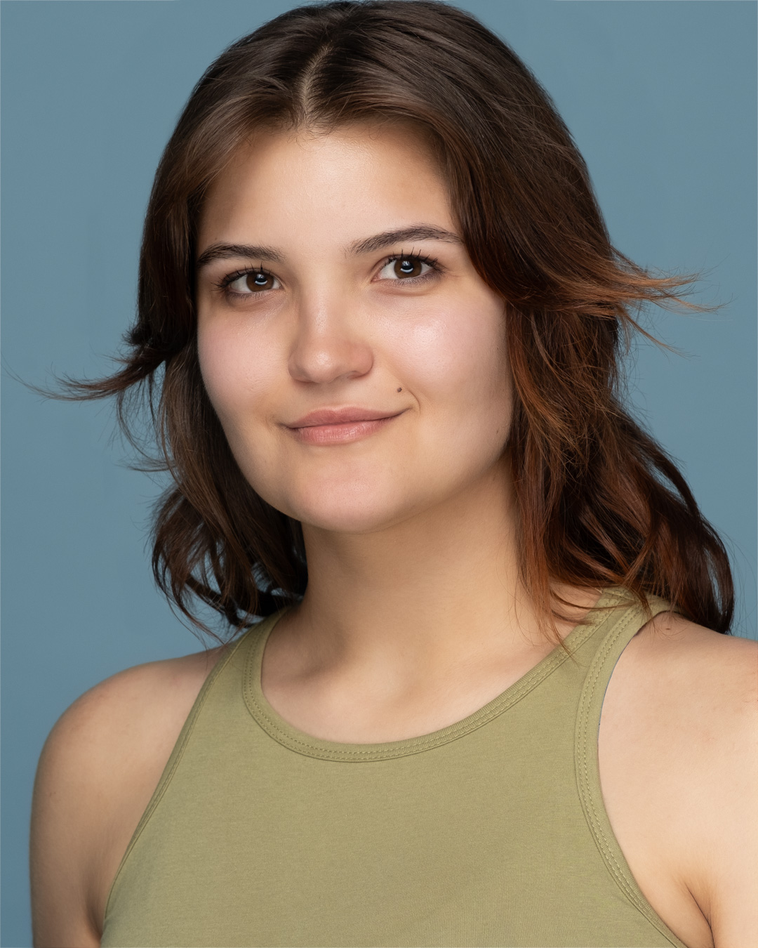 A young woman with shoulder-length brown hair wearing a light green sleeveless top poses against a plain blue background.