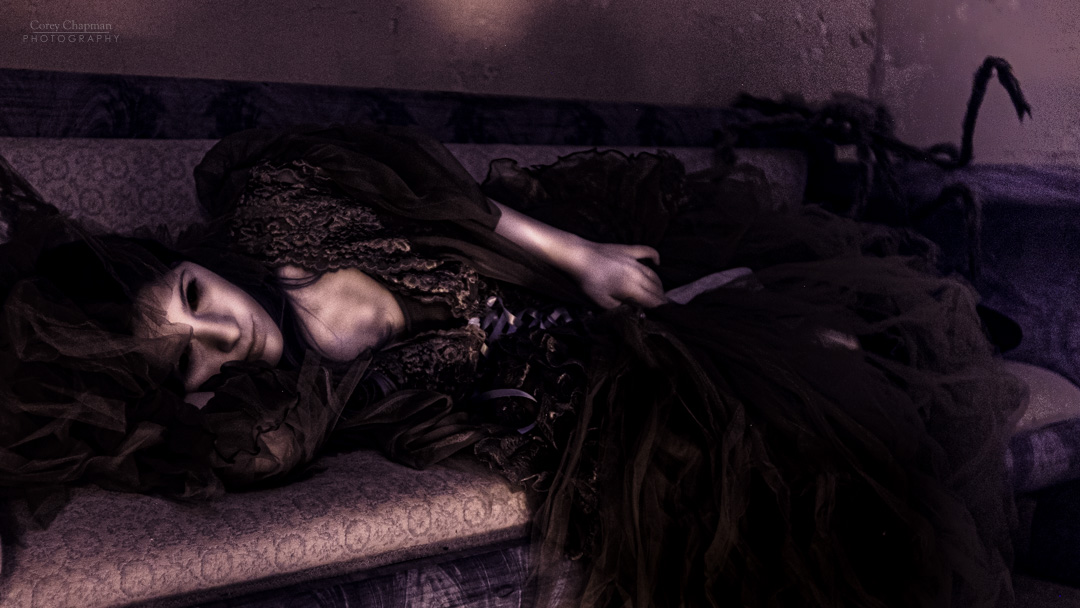 A person in dark, elaborate clothing lies on a sofa in a dimly lit room.