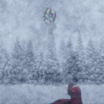 Person in red dress sits on snow in a forest during a snowstorm. A tall structure with colorful decorations is faintly visible in the background.