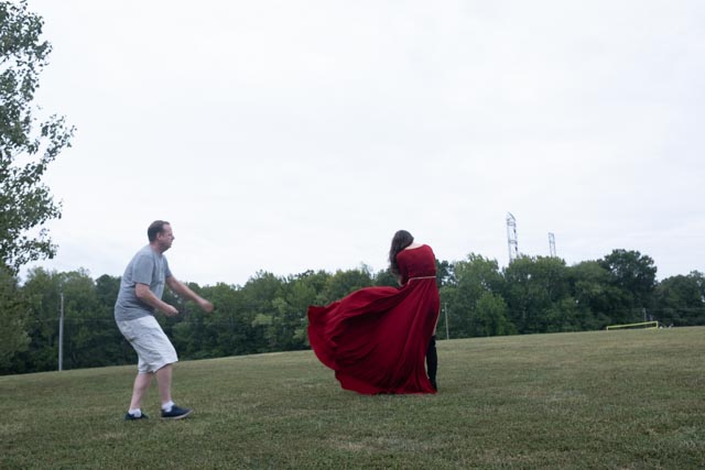 A man in shorts approaches a single girl in a flowing red dress standing on a grassy field under an overcast sky, creating a scene as enchanting as Christmas.