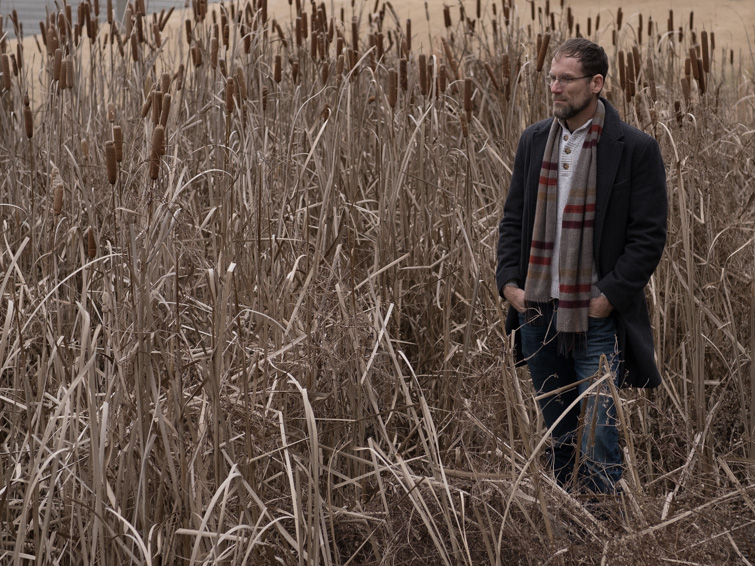 A person stands in a field of tall, dry reeds, wearing a dark coat and a plaid scarf.