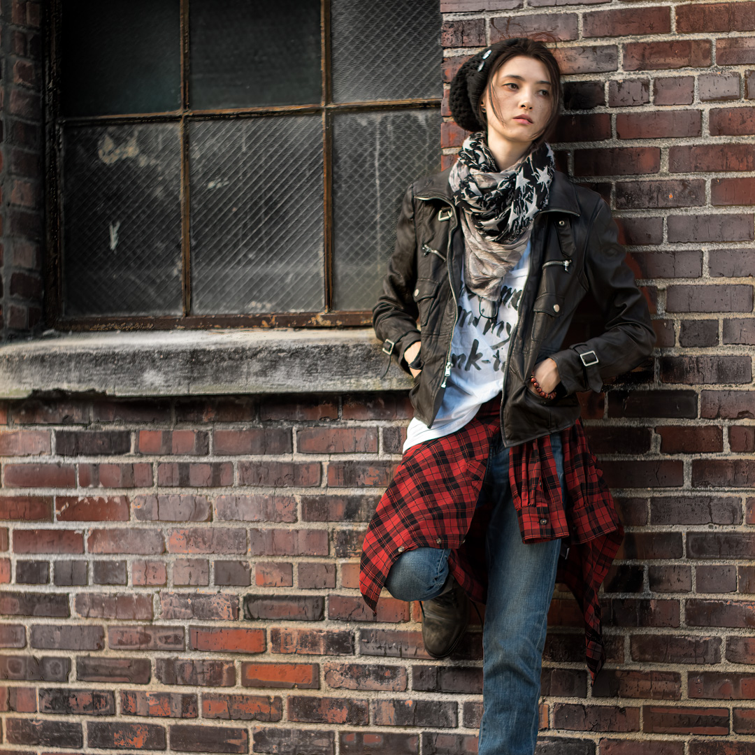 Person in casual attire, wearing a leather jacket, scarf, and jeans, leans against a brick wall with a window.