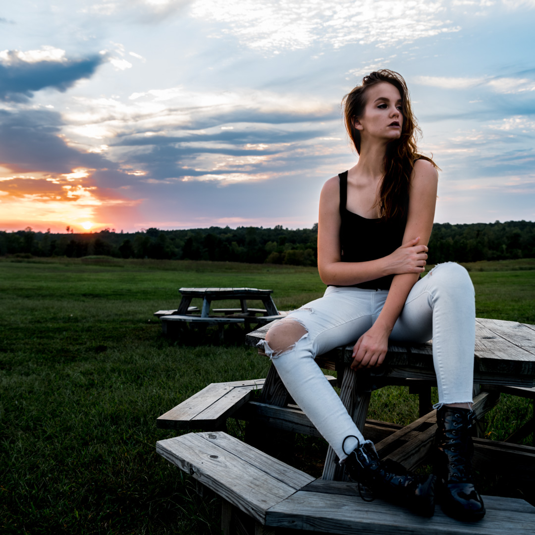 Person sitting on a wooden picnic table in a grassy field at sunset, wearing a black top, white ripped jeans, and black boots.