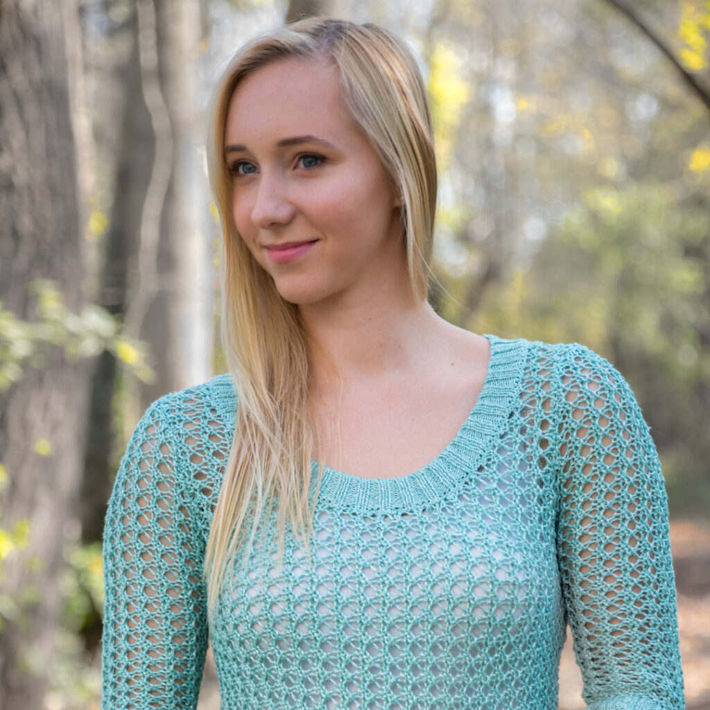 A person with long blonde hair wearing a light green knit sweater stands outdoors in a forested area.