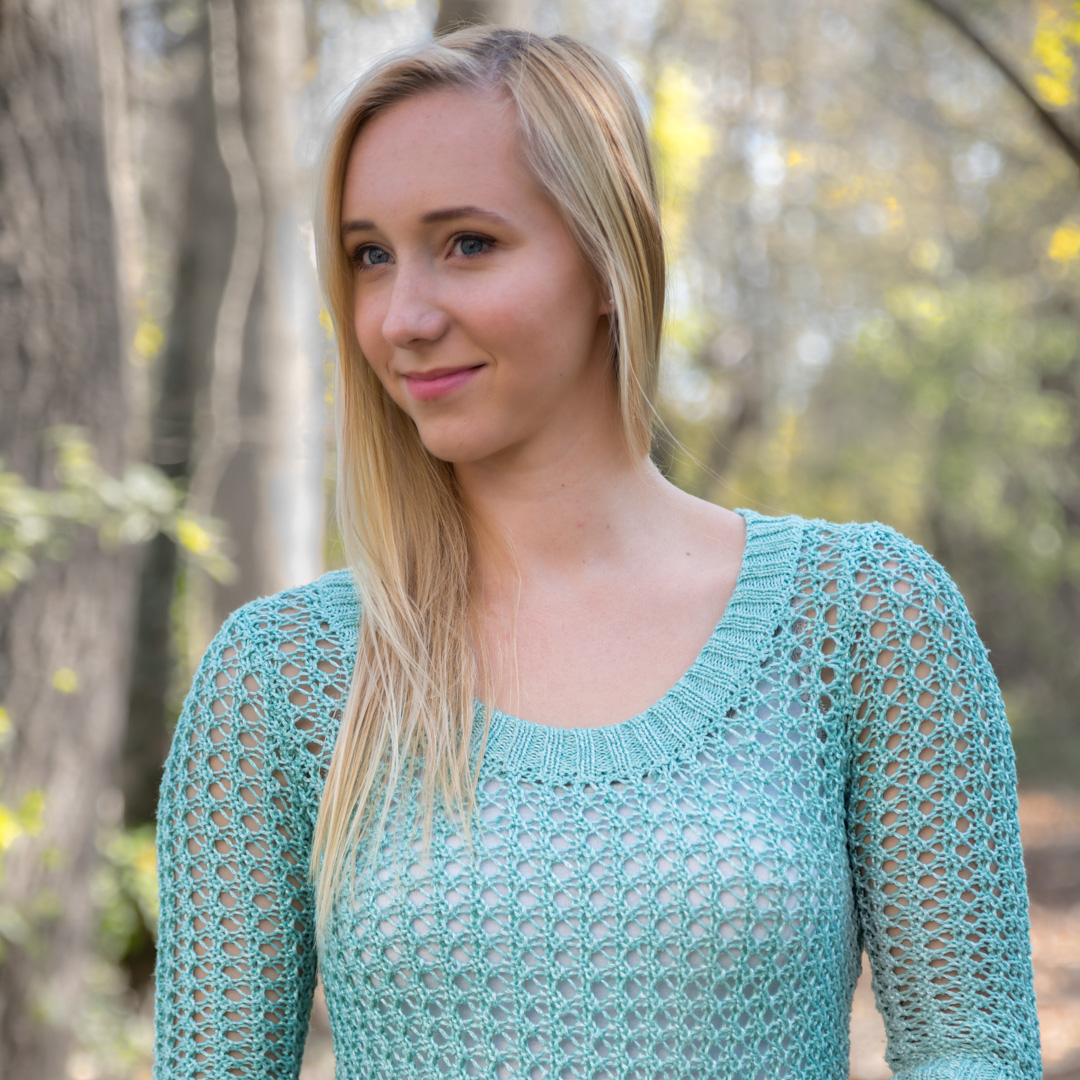 A person with long blonde hair wearing a light green knit sweater stands outdoors in a forested area.