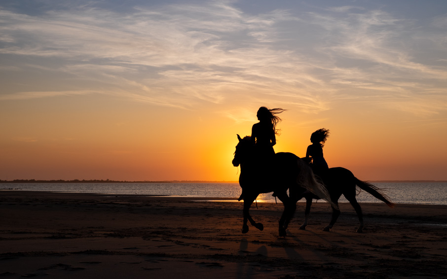 Two people ride horses along a beach at sunset, silhouetted against an orange sky and calm ocean.