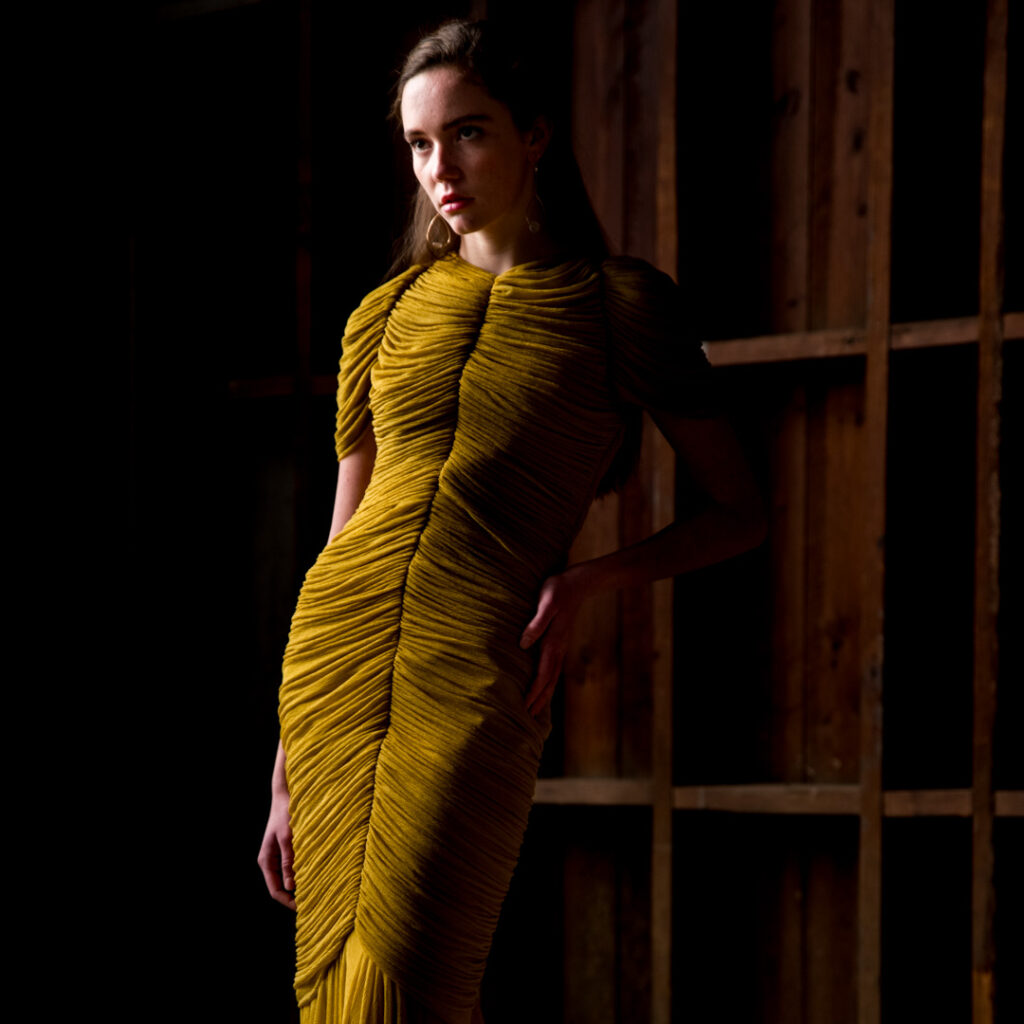 Person wearing a textured gold dress stands against a wooden backdrop, posing with one hand on hip in dramatic lighting.