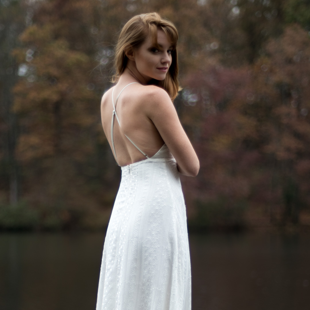 Woman in a white dress stands by a lake, looking over her shoulder. Background features blurred autumn trees.