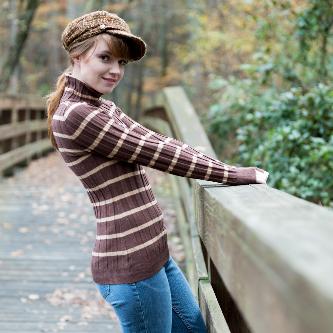 Person in a striped sweater and cap stands on a wooden bridge with trees in the background, slightly turned to the side.