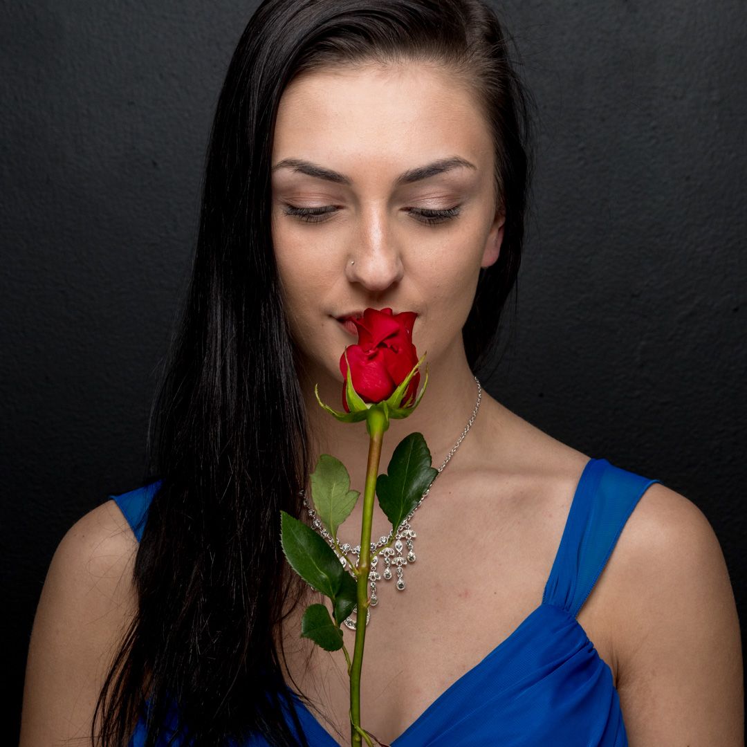 A woman with long dark hair holds a red rose near her face while wearing a blue dress and a necklace, set against a dark background.