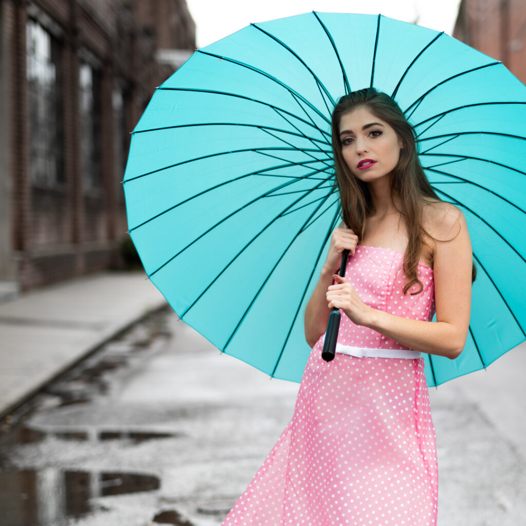 Woman in a pink polka dot dress stands on a rainy street holding a large turquoise umbrella.
